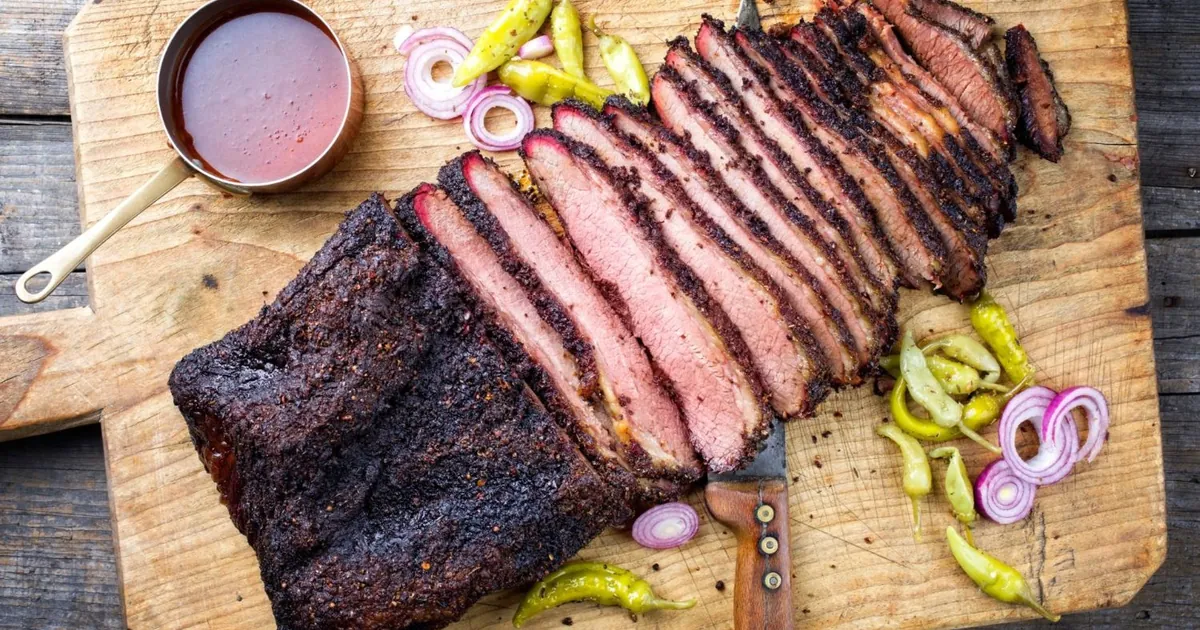 A sliced smoked brisket on a wooden board with a visible smoke ring, beside a small pot of sauce and pickled peppers