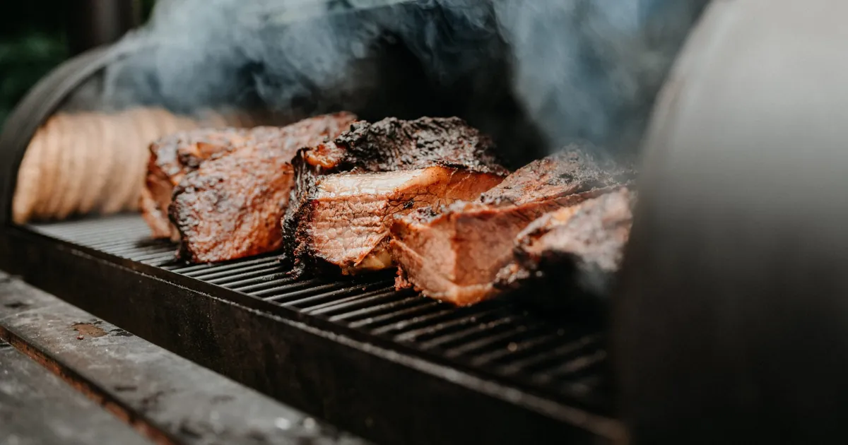 Brisket pieces with dark bark on the grate of an offset smoker, smoke rising under the open lid